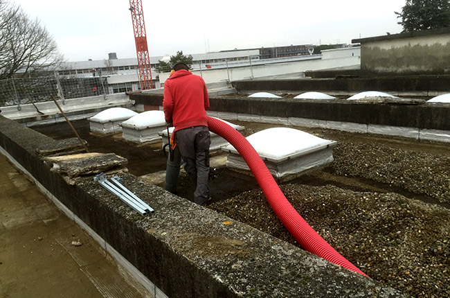 Rénovation de toit plat : Aspiration rapide du ballast. Rénovation de toit plat : aspiration du ballast par un ouvrier en sécurité, tuyau rouge, près de lanterneaux.