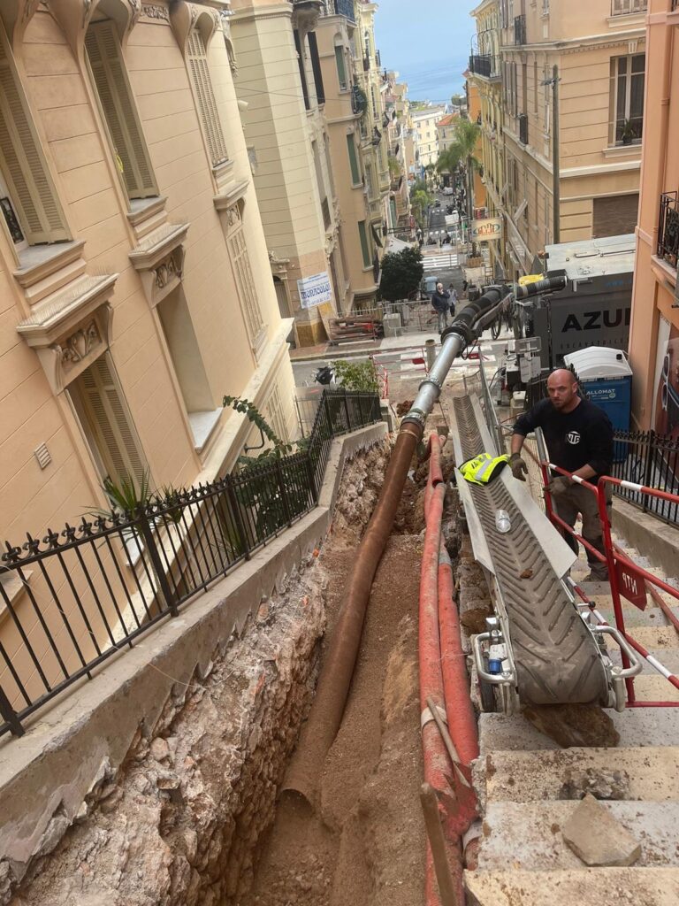 Construction à Monaco: Excavation dans un escalier raide. Travaux d'excavation sur un escalier raide de Monaco. Un ouvrier près d'un convoyeur retire la terre d'une tranchée profonde contenant des tuyaux. Vue sur la mer.