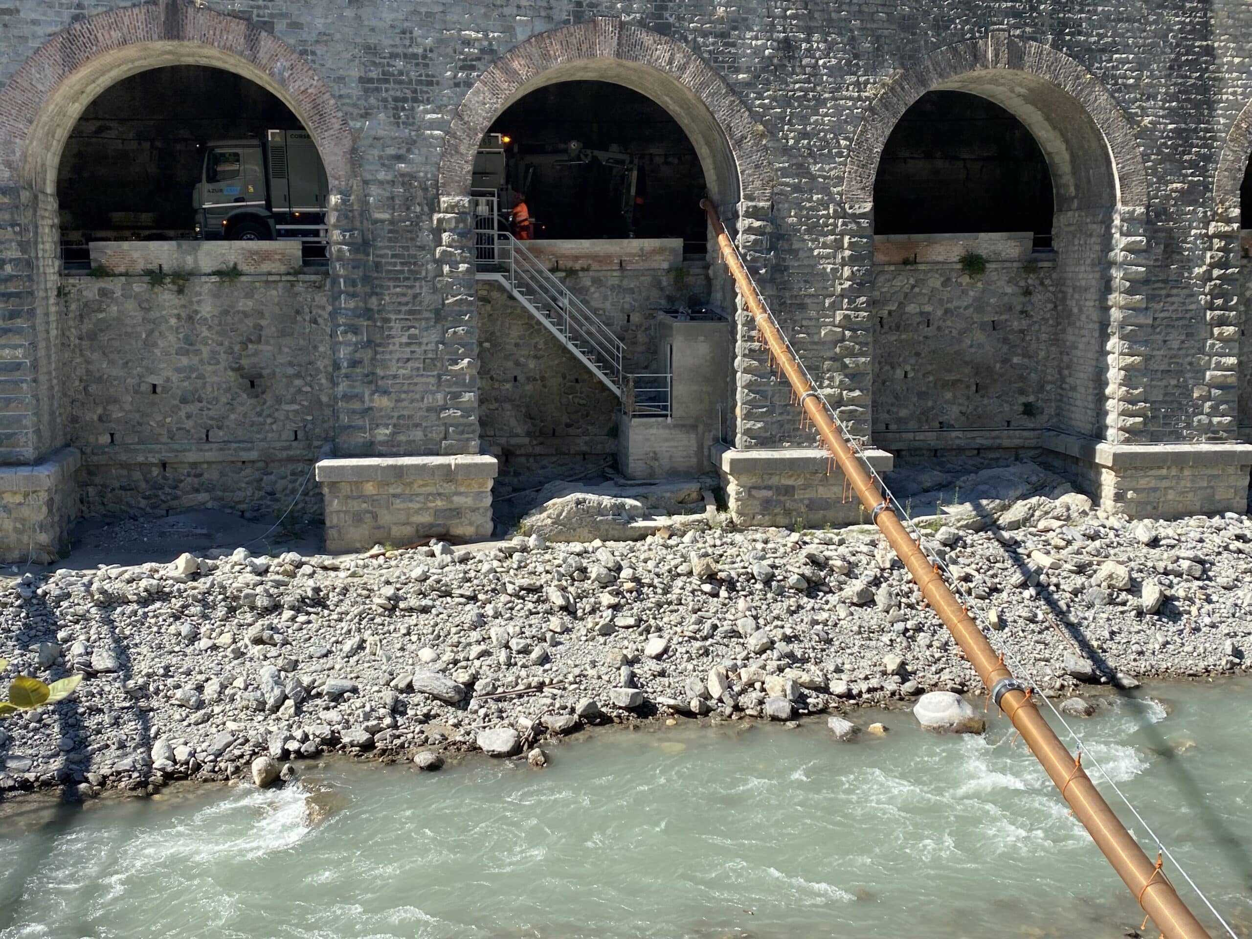 Arches d'un pont en pierre sur une rivière, travaux avec camion Azur et long tuyau orange sur le lit de galets.