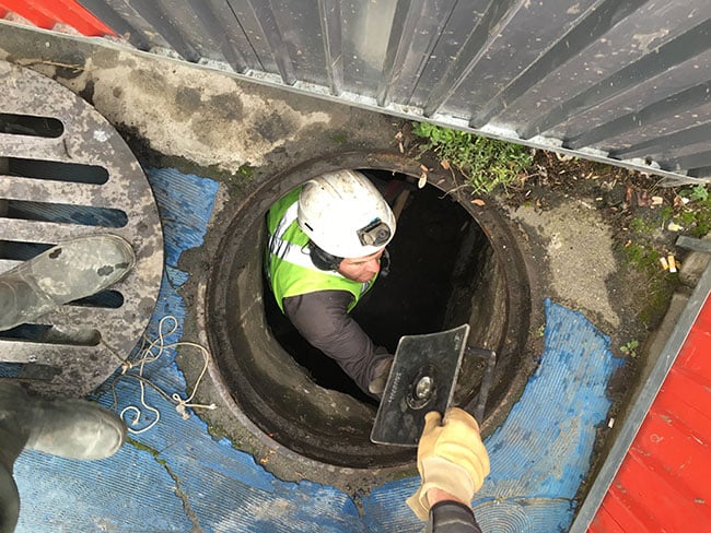 Travaux en espace confiné : entretien d'un regard d'égout. Ouvrier en casque et gilet fluorescent descendant dans un regard d'égout étroit. Travaux d'entretien en espace confiné.