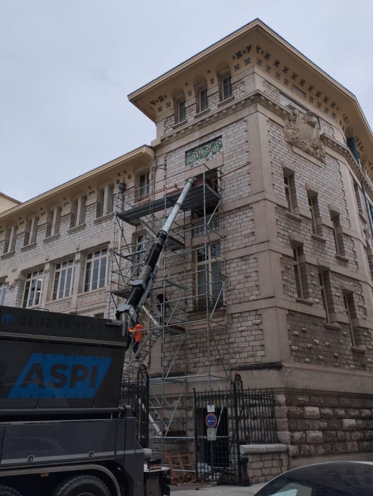 Rénovation de façade avec camion d'aspiration ASPI Camion ASPI aspirant des gravats via un bras articulé sur un échafaudage devant une façade de bâtiment ancien.