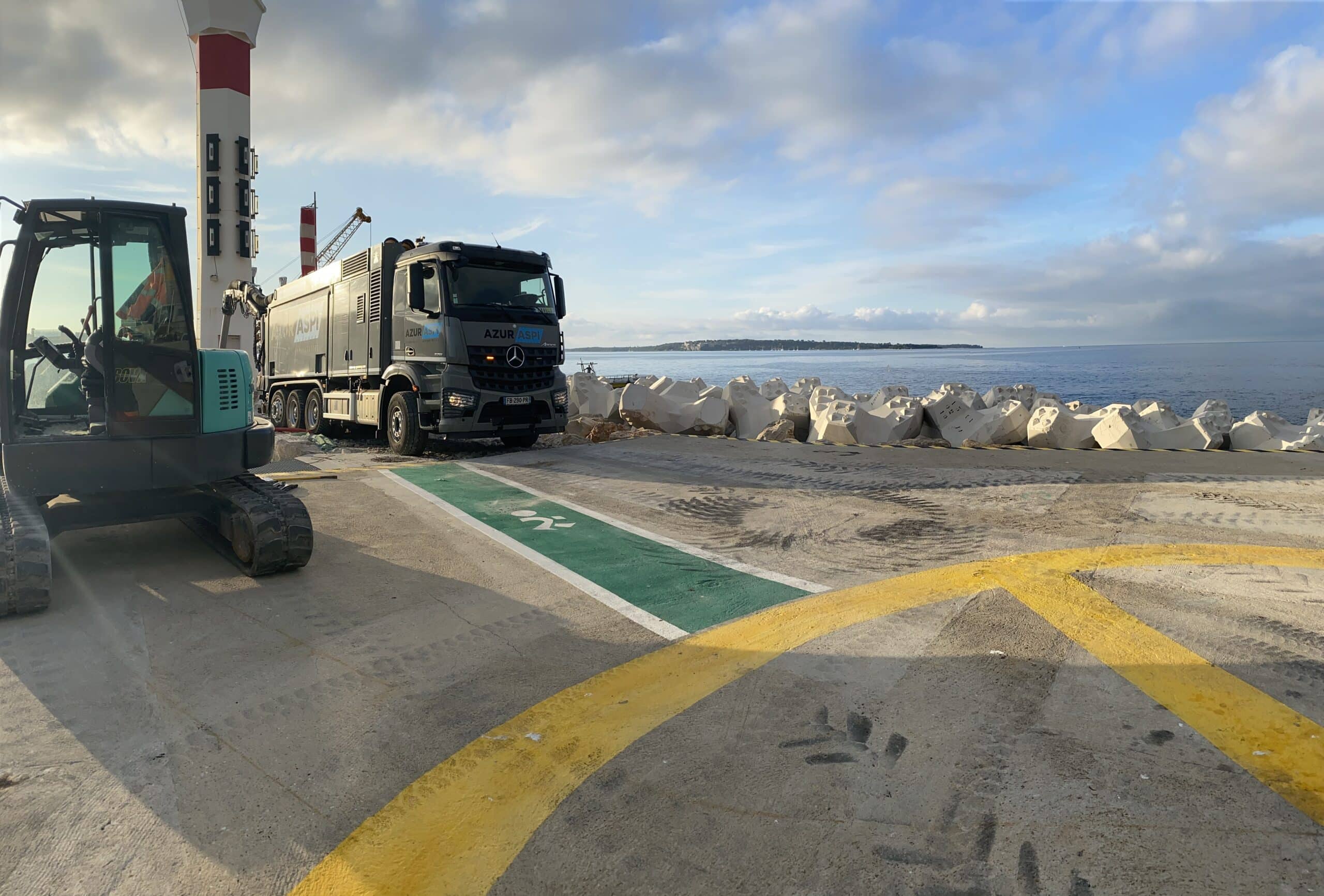 Camion aspirateur Azur Aspi et mini-pelle sur un chantier maritime face aux blocs de protection côtière.
