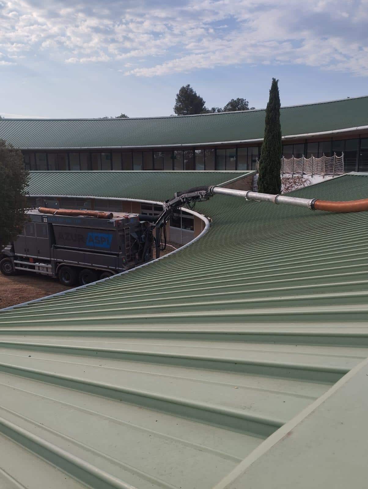 Camion aspirateur Azur Aspi avec bras articulé sur une toiture en métal vert incurvée d'un bâtiment moderne.