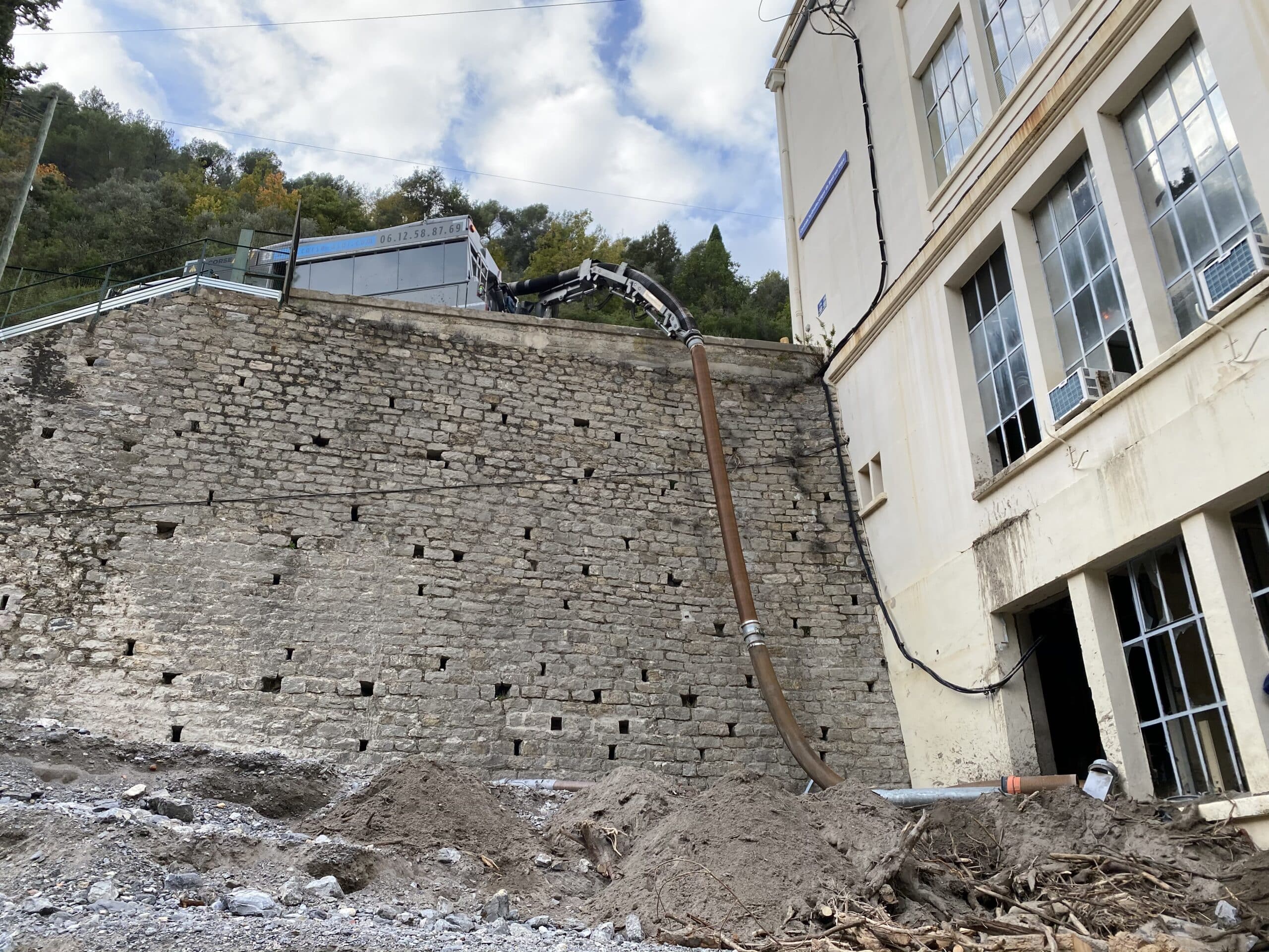 Camion aspirateur Corse Aspiration déblayant un chantier contre un mur en pierre avec un long tuyau flexible.