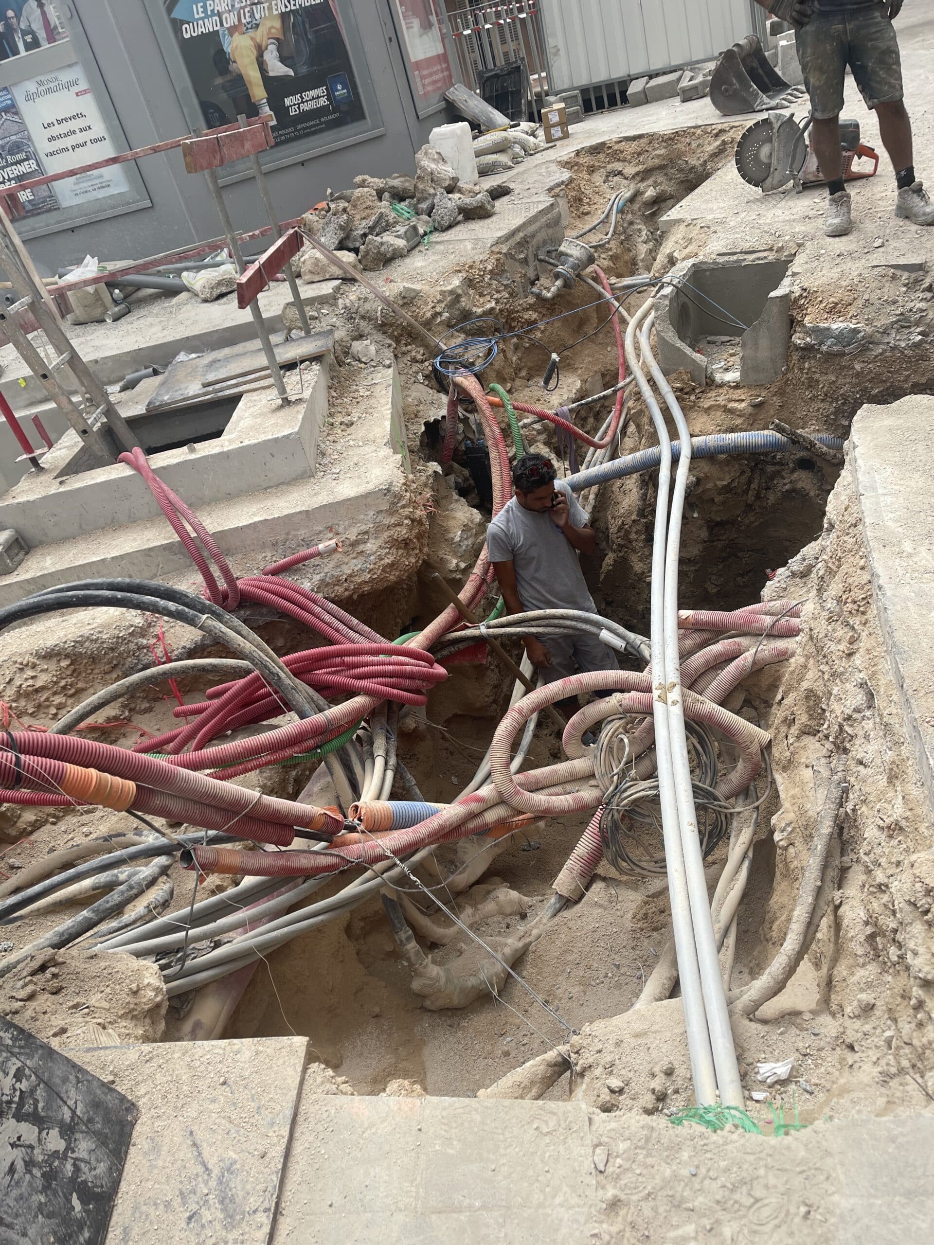 Ouvrier dans une tranchée de chantier urbain au milieu de nombreux câbles, tuyaux et gaines de protection rouges.