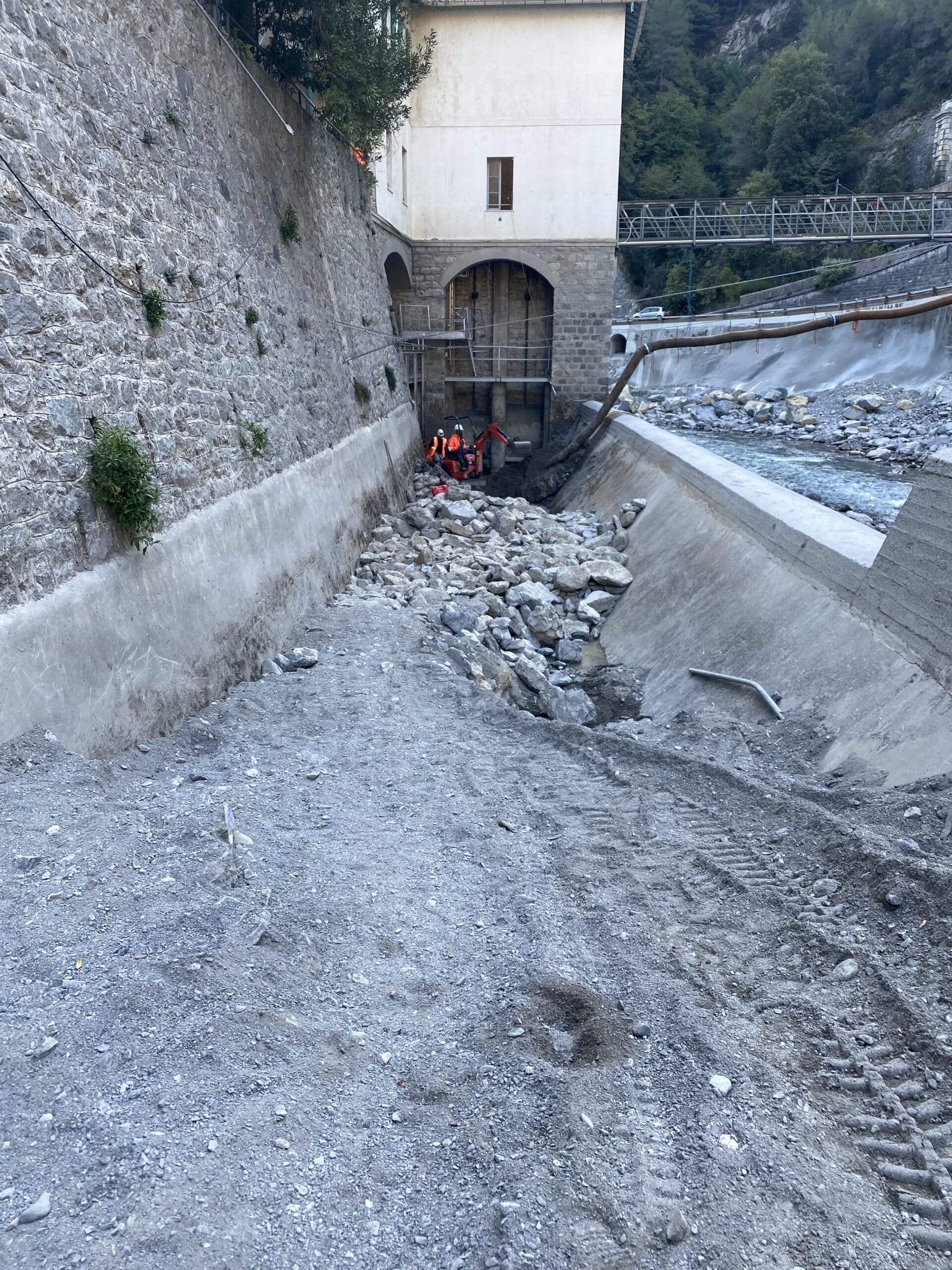 Ouvriers et mini-pelle en plein chantier de renforcement des berges rocheuses près d'un cours d'eau.