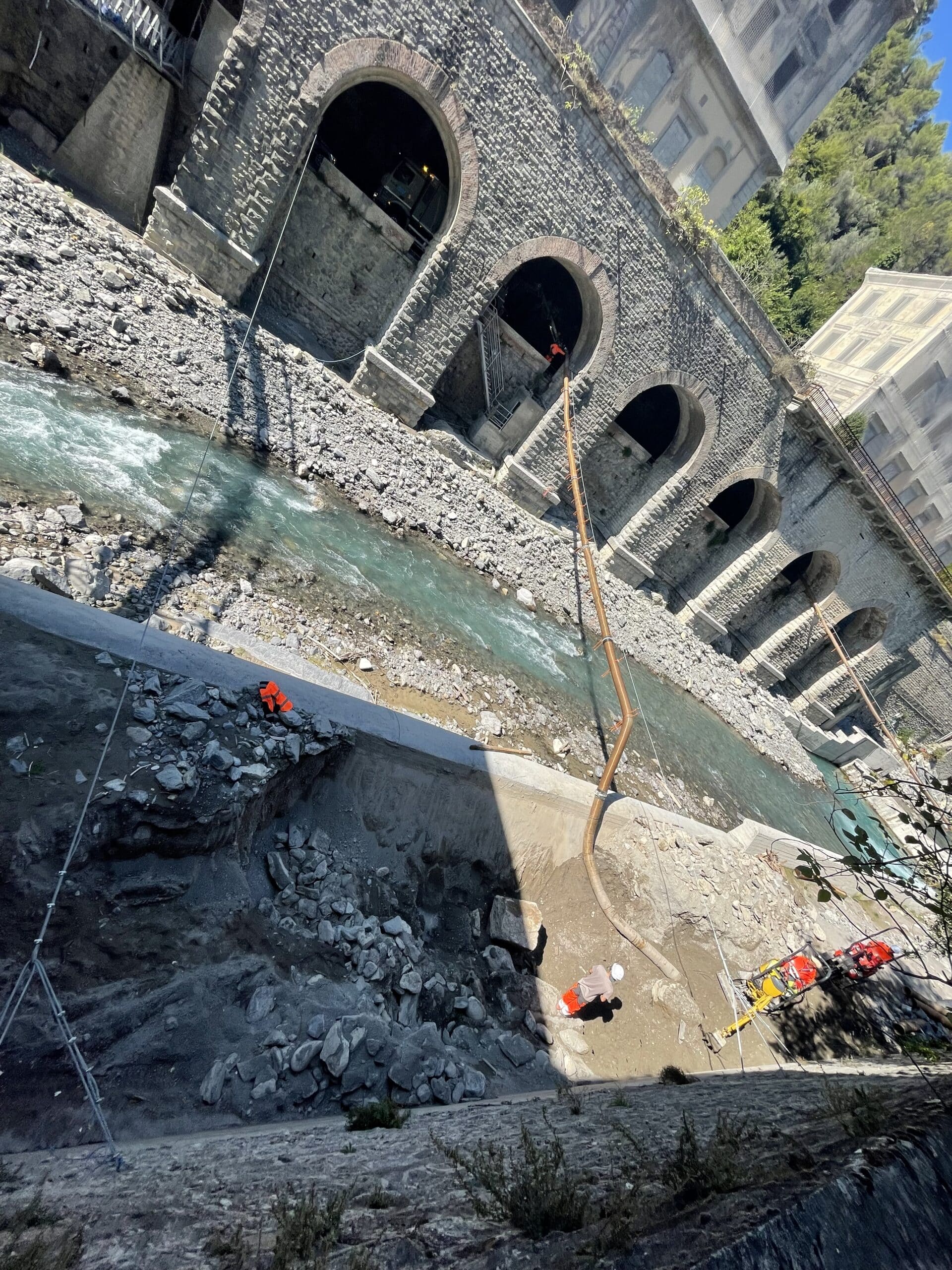 Chantier de restauration sous des arches en pierre en bord de rivière avec ouvriers et pelleteuse.