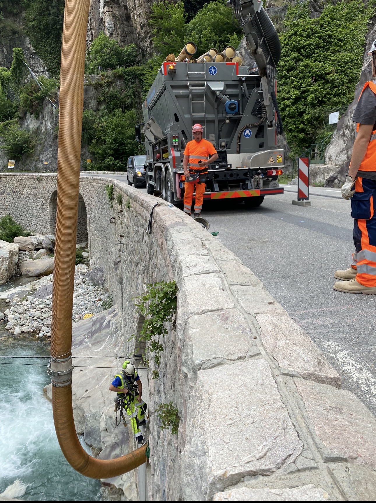 Ouvrier en rappel et camion aspirateur effectuant la maintenance d'un pont en pierre au-dessus d'une rivière de montagne.