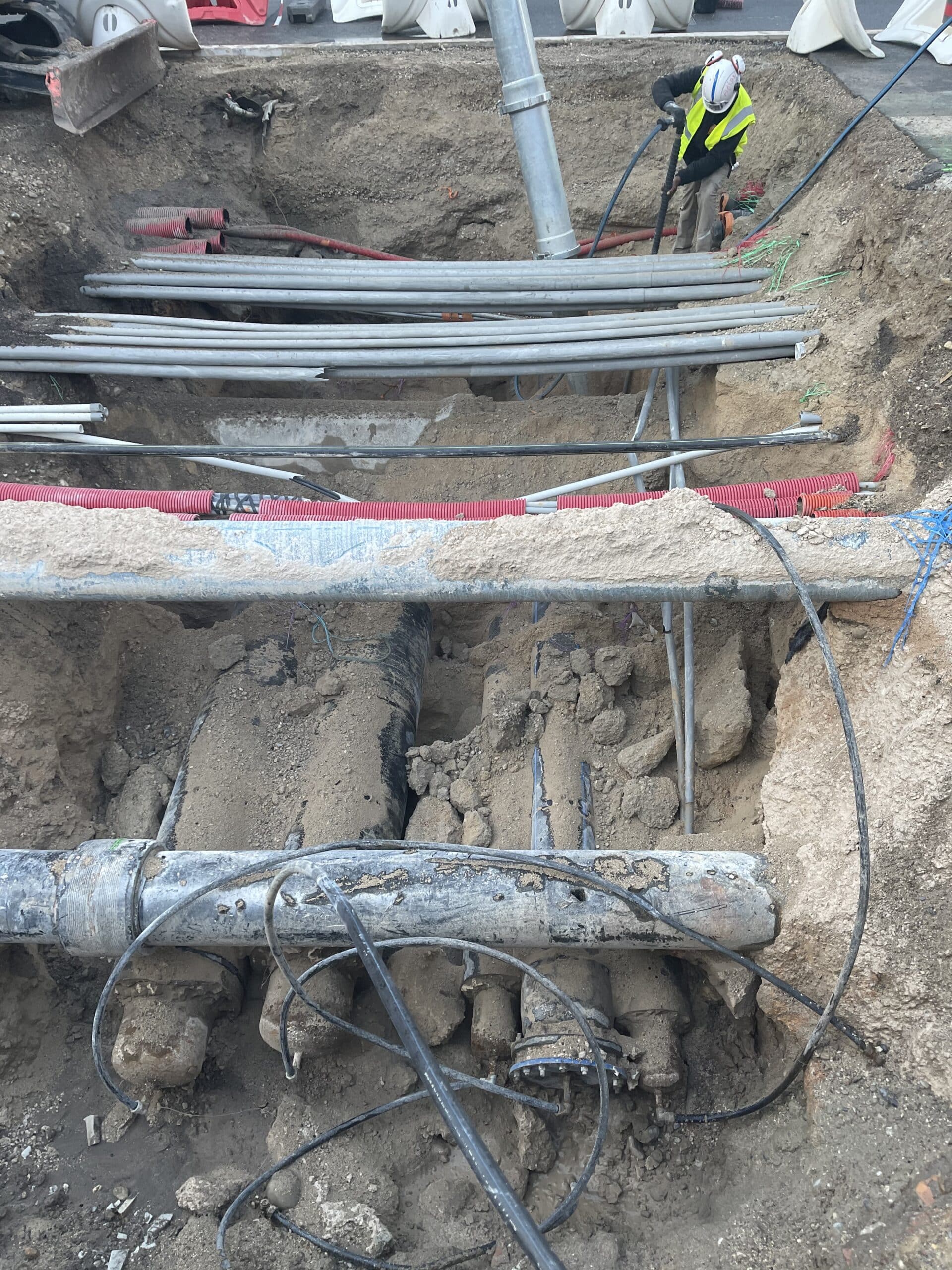 Ouvrier au travail dans une tranchée révélant un réseau dense de tuyaux, câbles et canalisations souterraines.