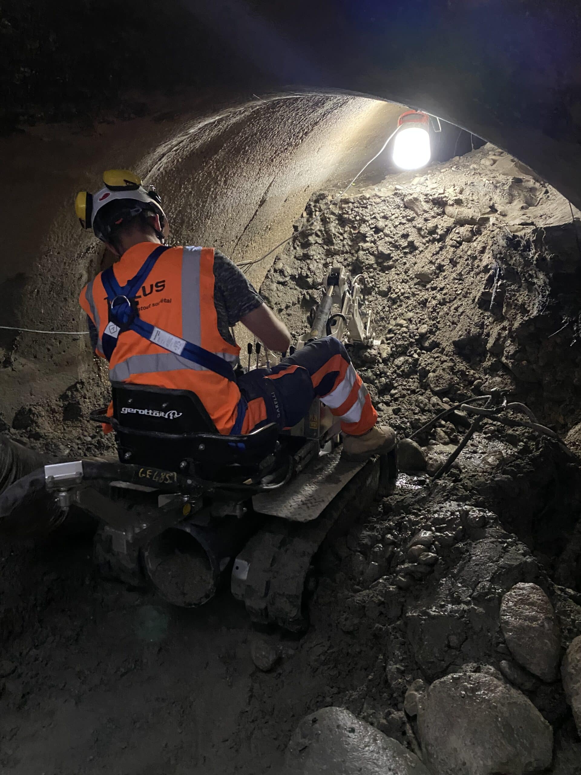 Ouvrier Telus opérant une mini-pelle dans un tunnel sombre, avec gilet de sécurité orange et harnais.