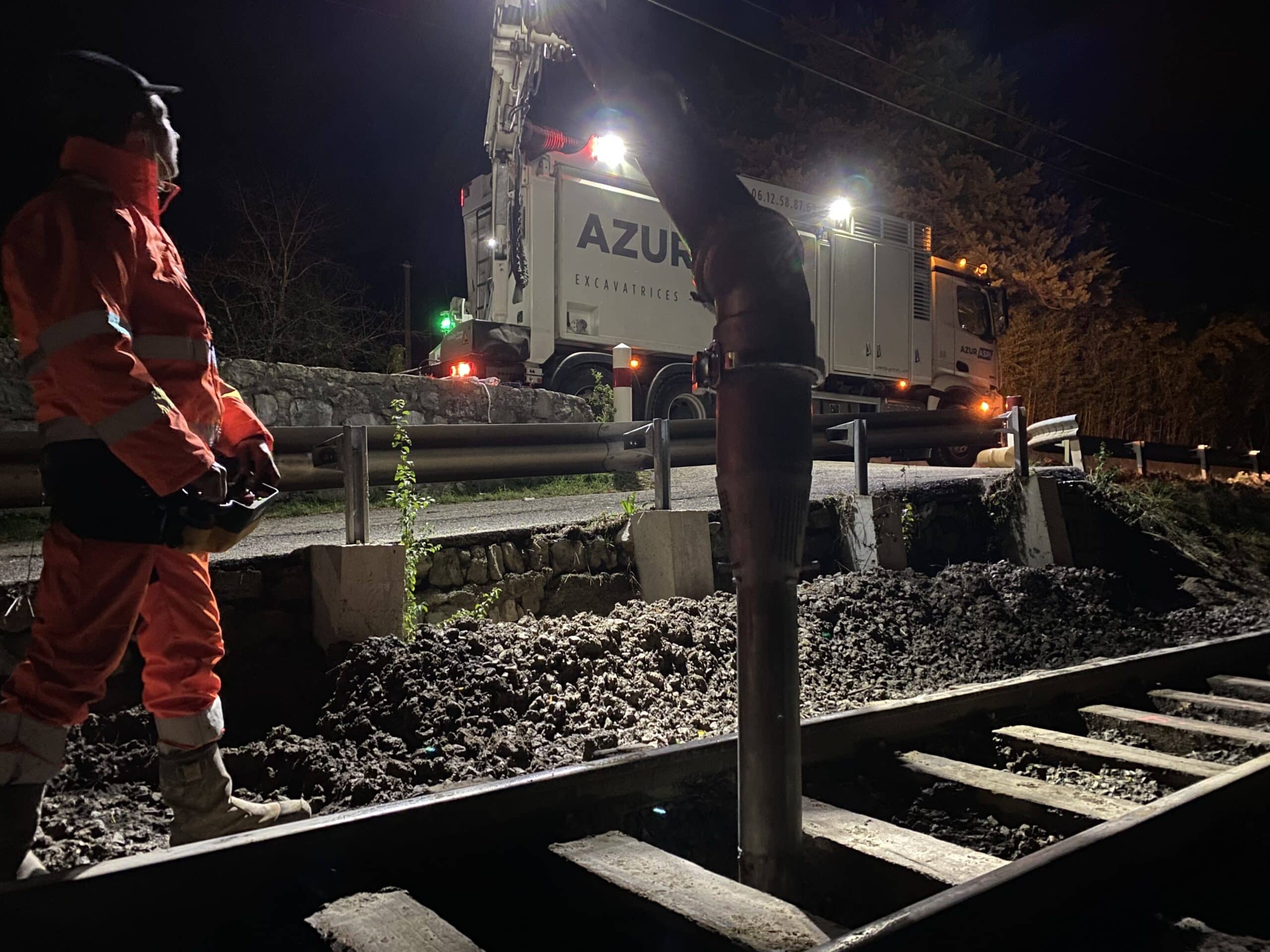 Ouvrier et excavatrice aspiratrice Azur effectuant des travaux ferroviaires nocturnes sur une voie ferrée décaissée.