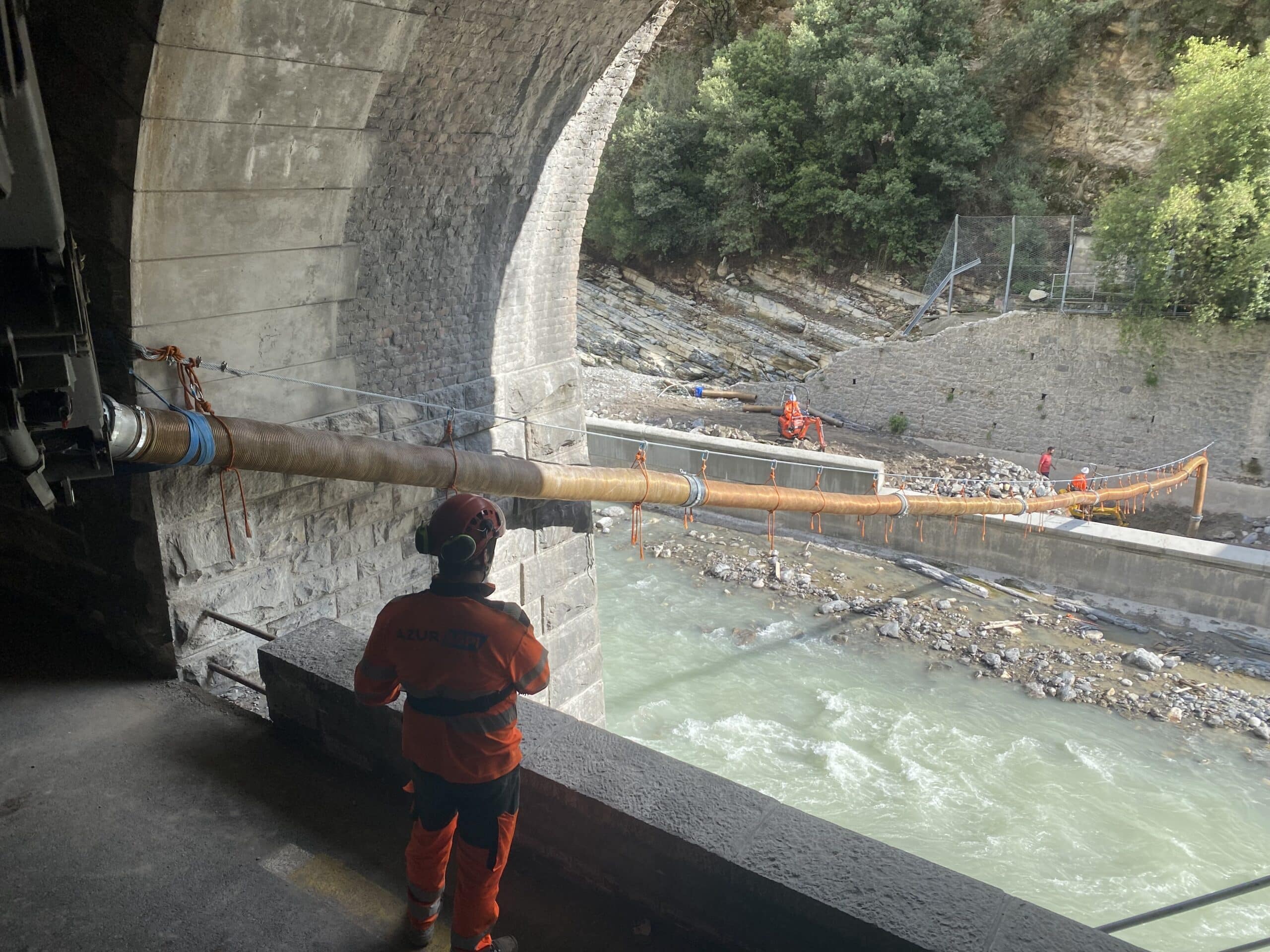 Ouvrier Azur ASPT surveillant un tuyau suspendu au-dessus d'une rivière depuis un tunnel en pierre.