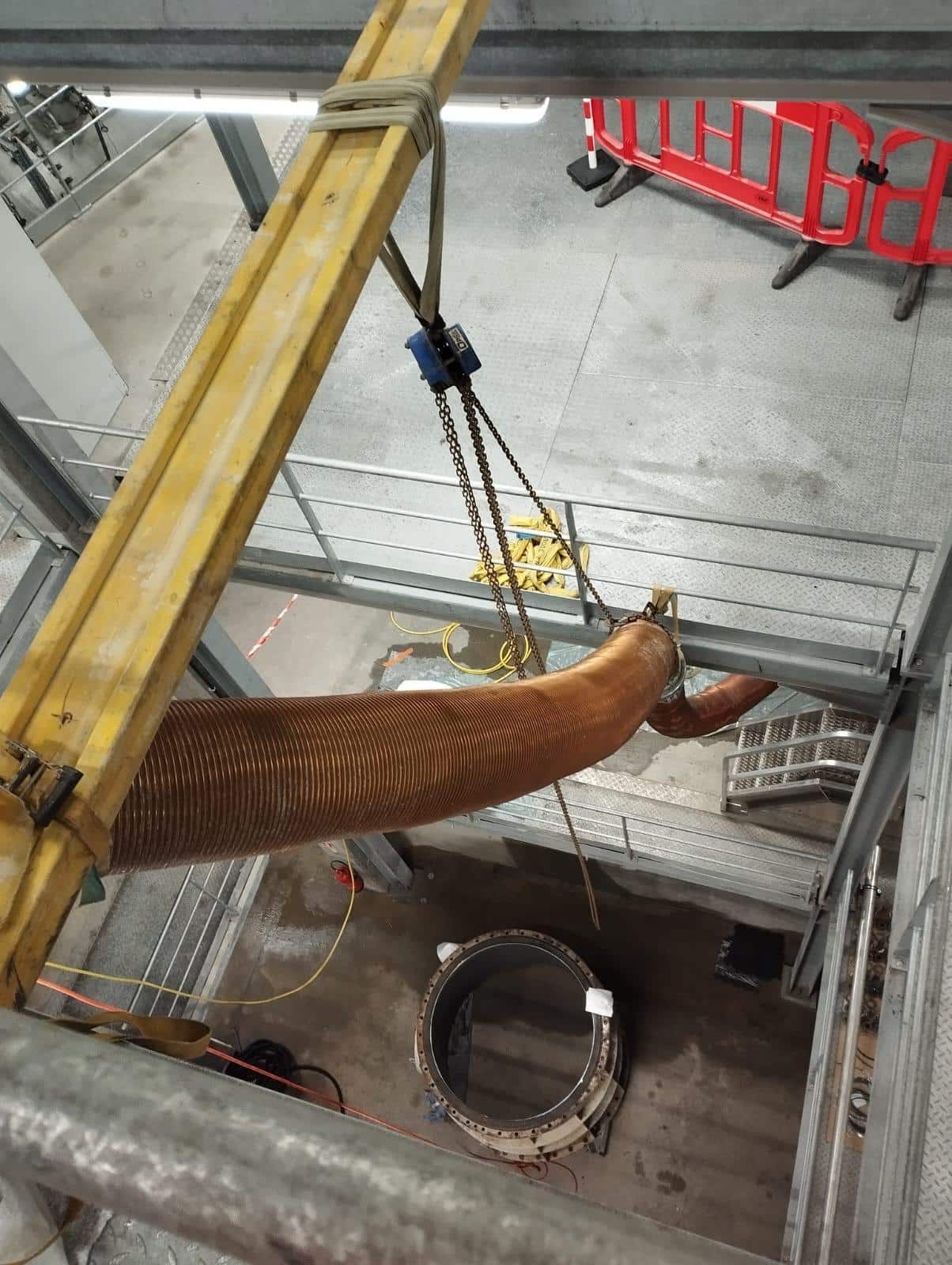 Levage d'un conduit flexible orange avec palan à chaîne au-dessus d'une bride métallique en milieu industriel.