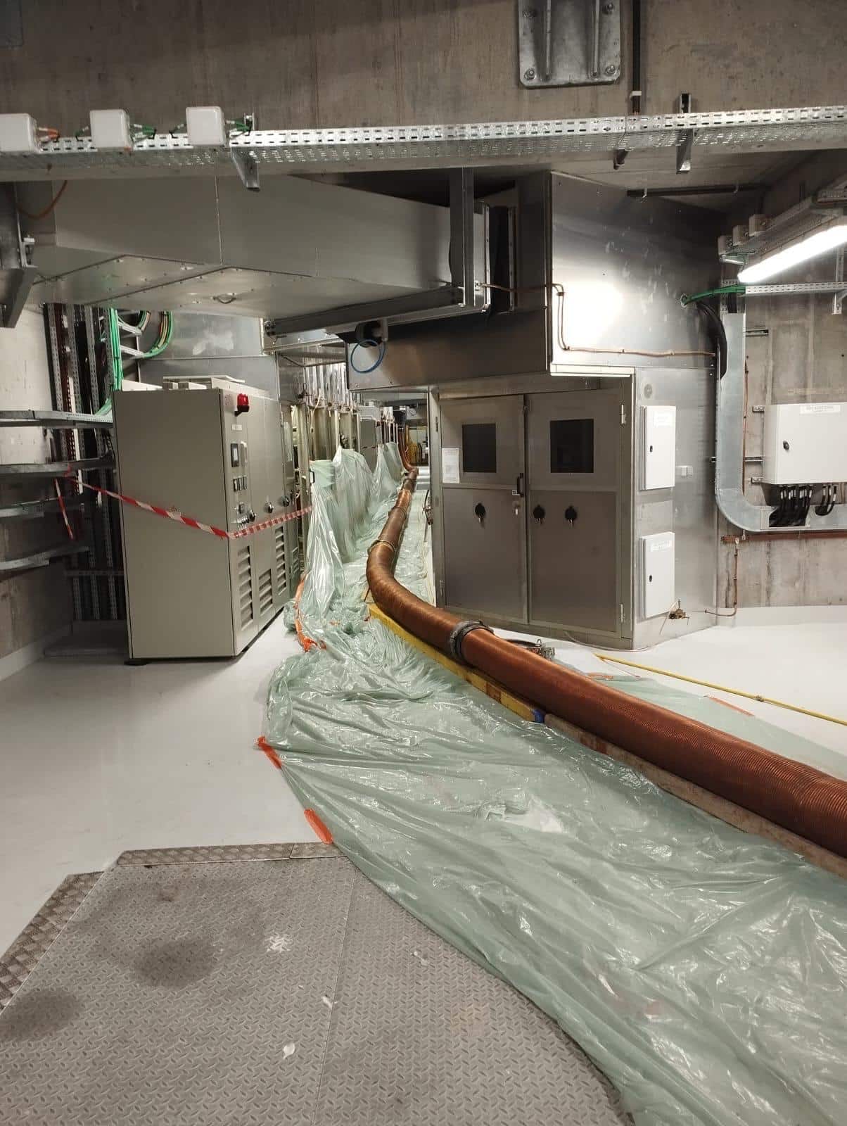 Maintenance industrielle d'une salle technique avec tuyau flexible, bâches plastiques et armoires électriques.
