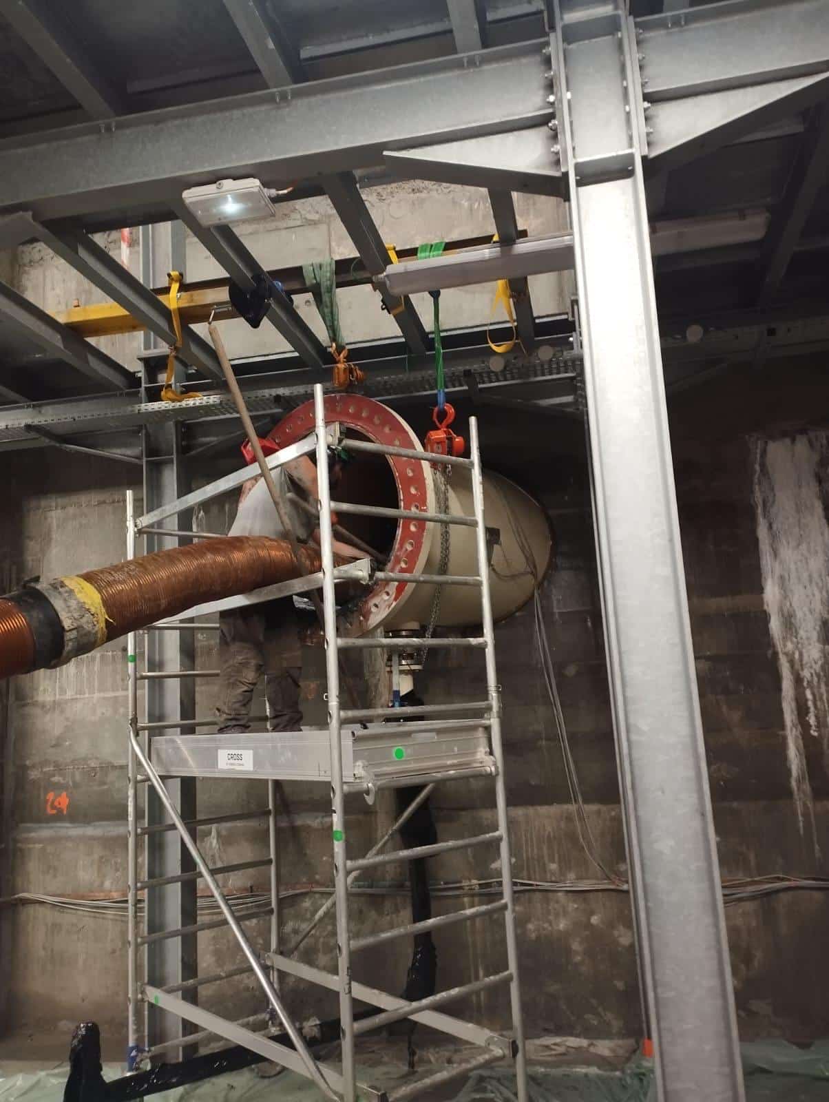 Ouvrier sur échafaudage travaillant sur une conduite industrielle avec bride rouge et câble flexible.