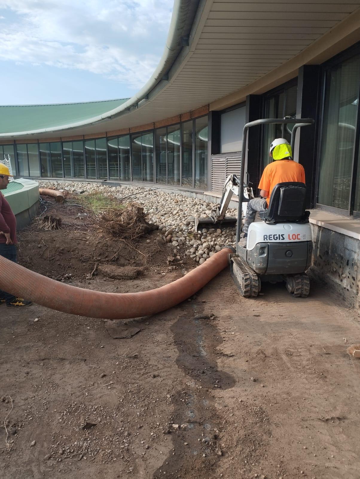 Mini-pelle REGIS LOC déplaçant des galets sur un chantier de terrassement près d'un bâtiment vitré moderne.