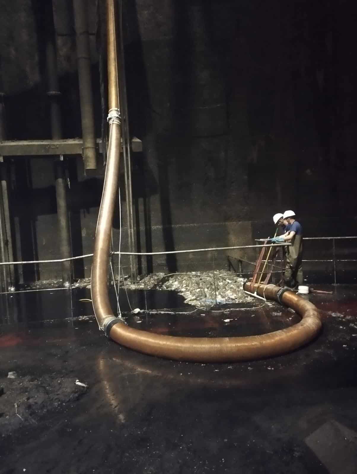 Ouvriers industriels manipulant un large tuyau d'aspiration pour nettoyer des sédiments dans un bassin sombre.