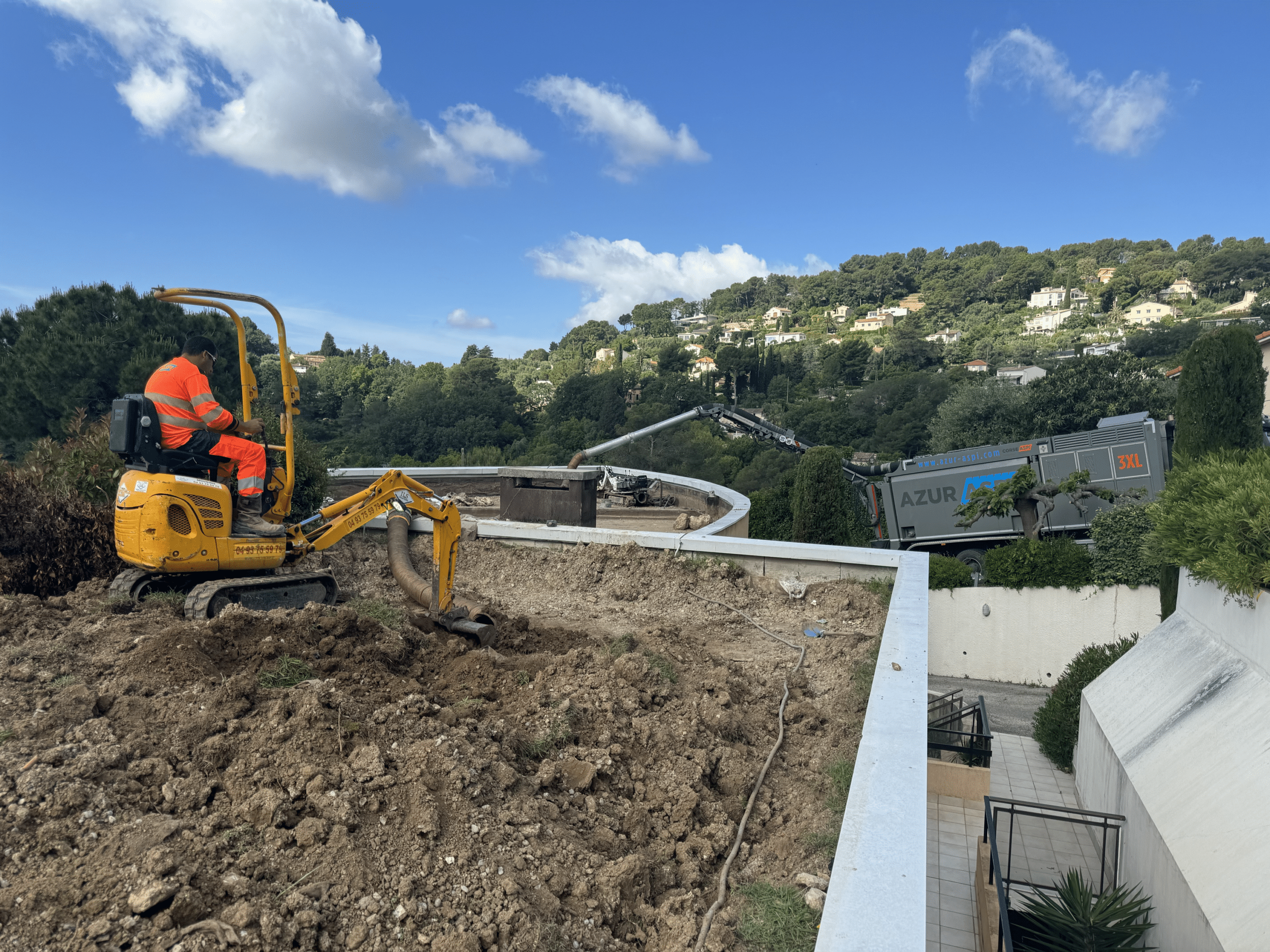 Ouvrier utilisant une mini-pelle jaune et un camion aspirateur Azur Aspi 3XL pour un terrassement de jardin en pente.