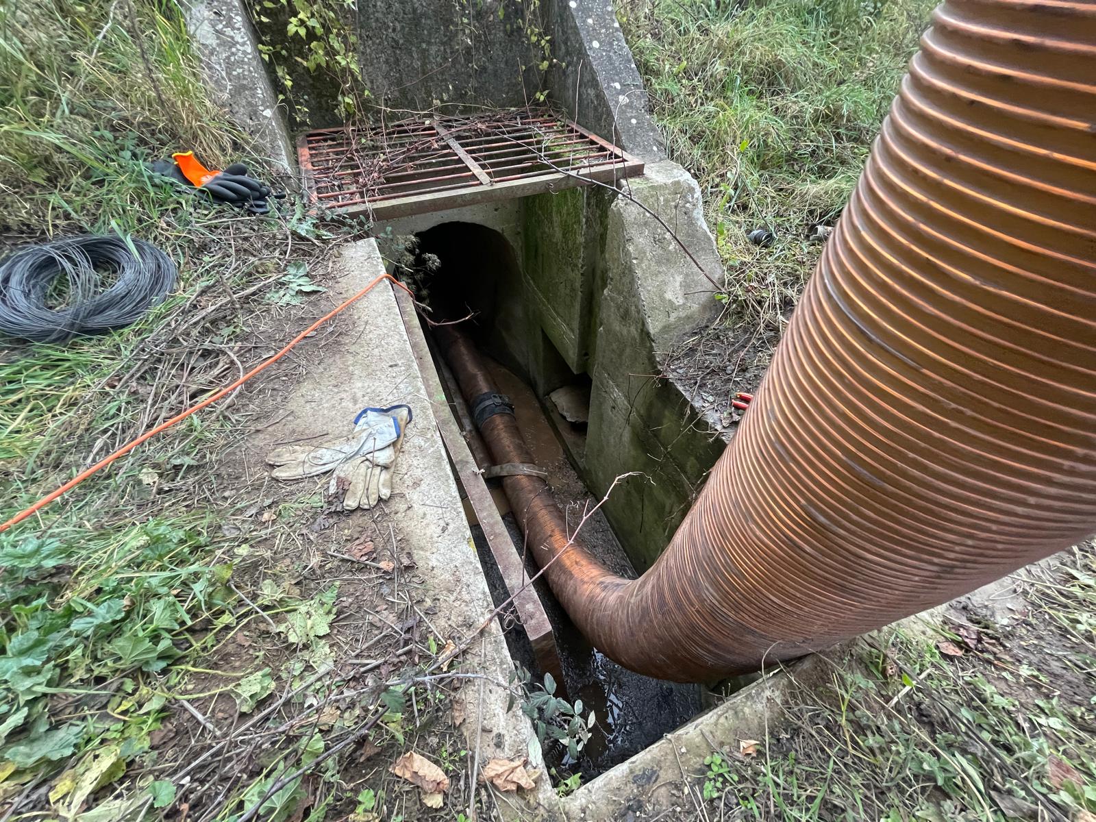 Tuyau annelé orange inséré dans un ponceau en béton avec grille de fer et équipement de chantier au sol.